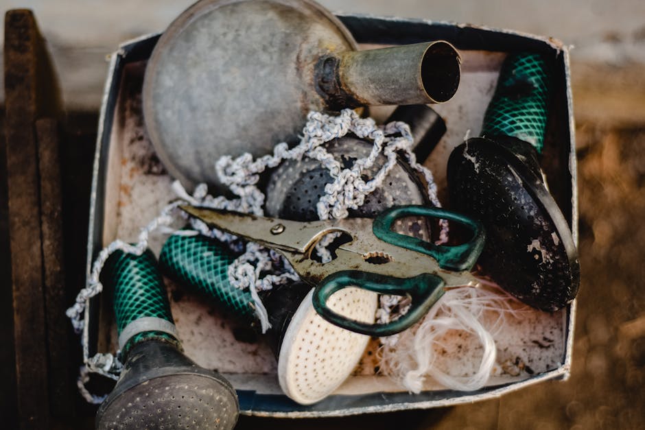 A close-up view of a metal toolbox filled with gardening tools and accessories, placed outdoors. Inside the box, there are rubber gloves with green and black checkered patterns, a pair of small sharp